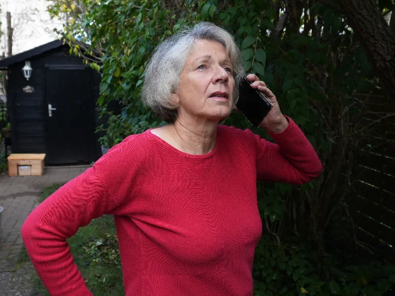 Older woman in a red sweater talking on a phone outdoors near trees and a shed.