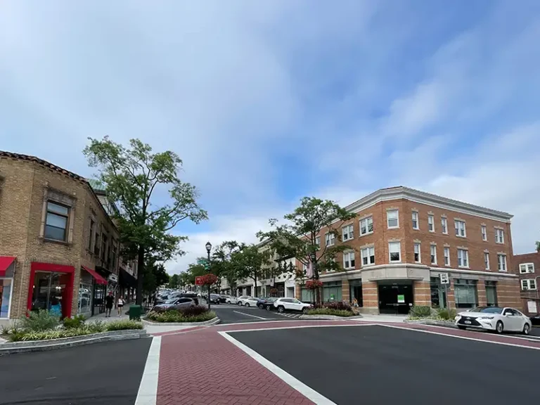 Downtown street with brick buildings, parked cars, and trees under a partly cloudy sky.