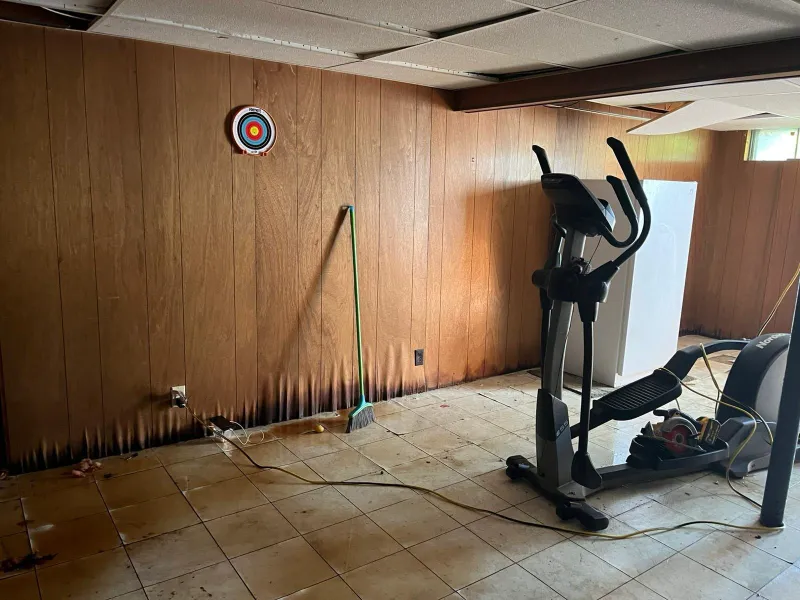 Elliptical machine and broom in a basement with wood-paneled walls and a small target on the wall.
