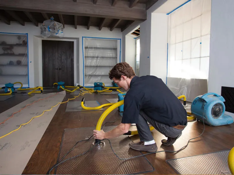 Man crouching while setting up yellow air ducts on a hardwood floor in a room under renovation.