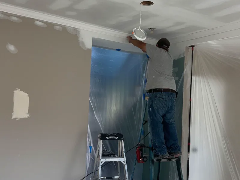 A man on a ladder installing or repairing a ceiling light fixture in a room under renovation.
