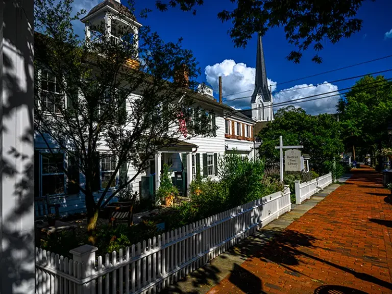 White colonial-style building with a picket fence and a church steeple in the background under a blue sky.