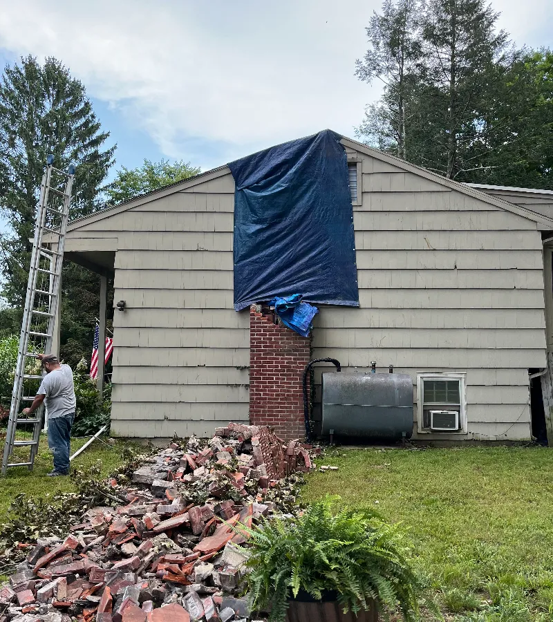 Man standing by a ladder next to a house with a damaged chimney covered by a tarp and bricks scattered on the ground.