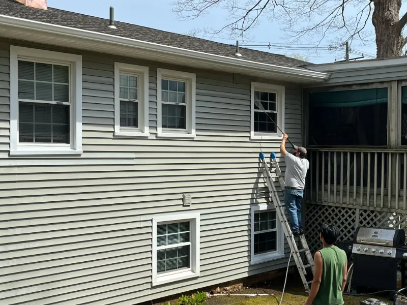 A man on a ladder painting the exterior siding of a house while another man watches.