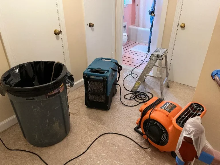 Dehumidifier and air mover equipment set up in a hallway outside a bathroom with a ladder and trash can.