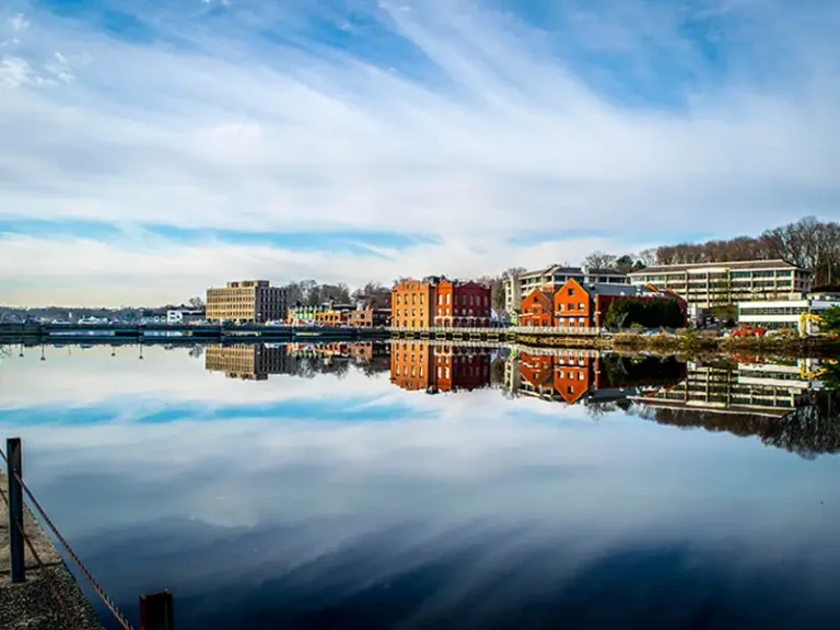 Buildings and trees reflected in calm river water under a partly cloudy sky.