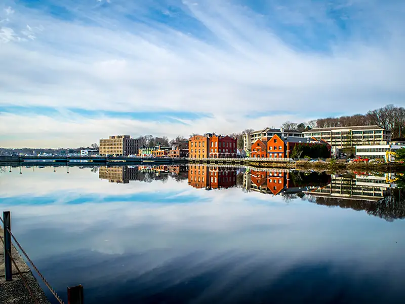 Buildings and trees reflected in calm river water under a partly cloudy sky.