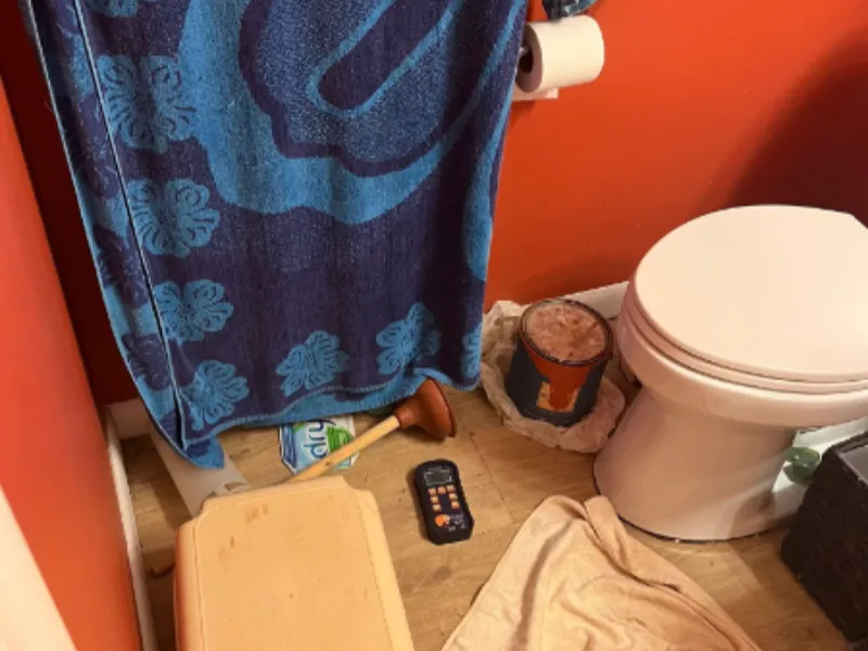 Bathroom floor with a toilet, plunger, remote control, and a blue towel hanging.
